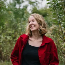 A woman with short blonde hair, wearing a red jacket over a black top, standing among trees.