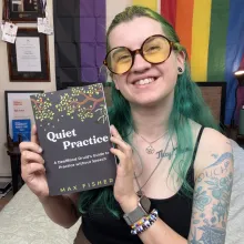 Max, a white genderqueer person with green hair, sat in front of a pride flag, holding a copy of their book. It is black with white text and green leaves on it. 