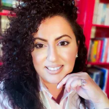 Headshot of author Joelle Habbaki with dark curly hair, resting her hand under her chin and looking directly at the camera, with a bright pink bookshelf in the background.